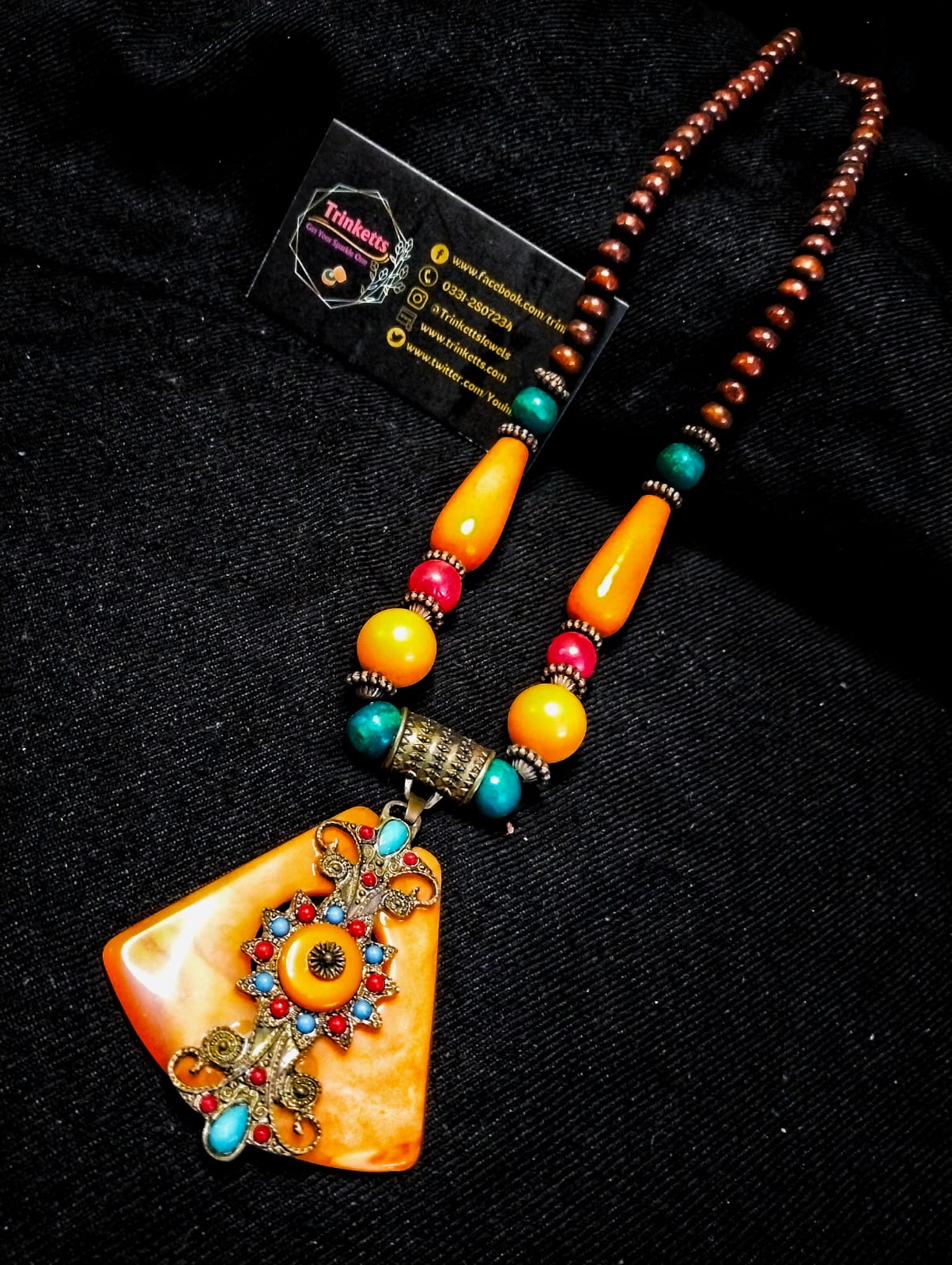 Triangular orange pendant with intricate bronze alloy design, accented by wooden and plastic beads in shades of brown, orange, and red, alongside elongated orange beads.