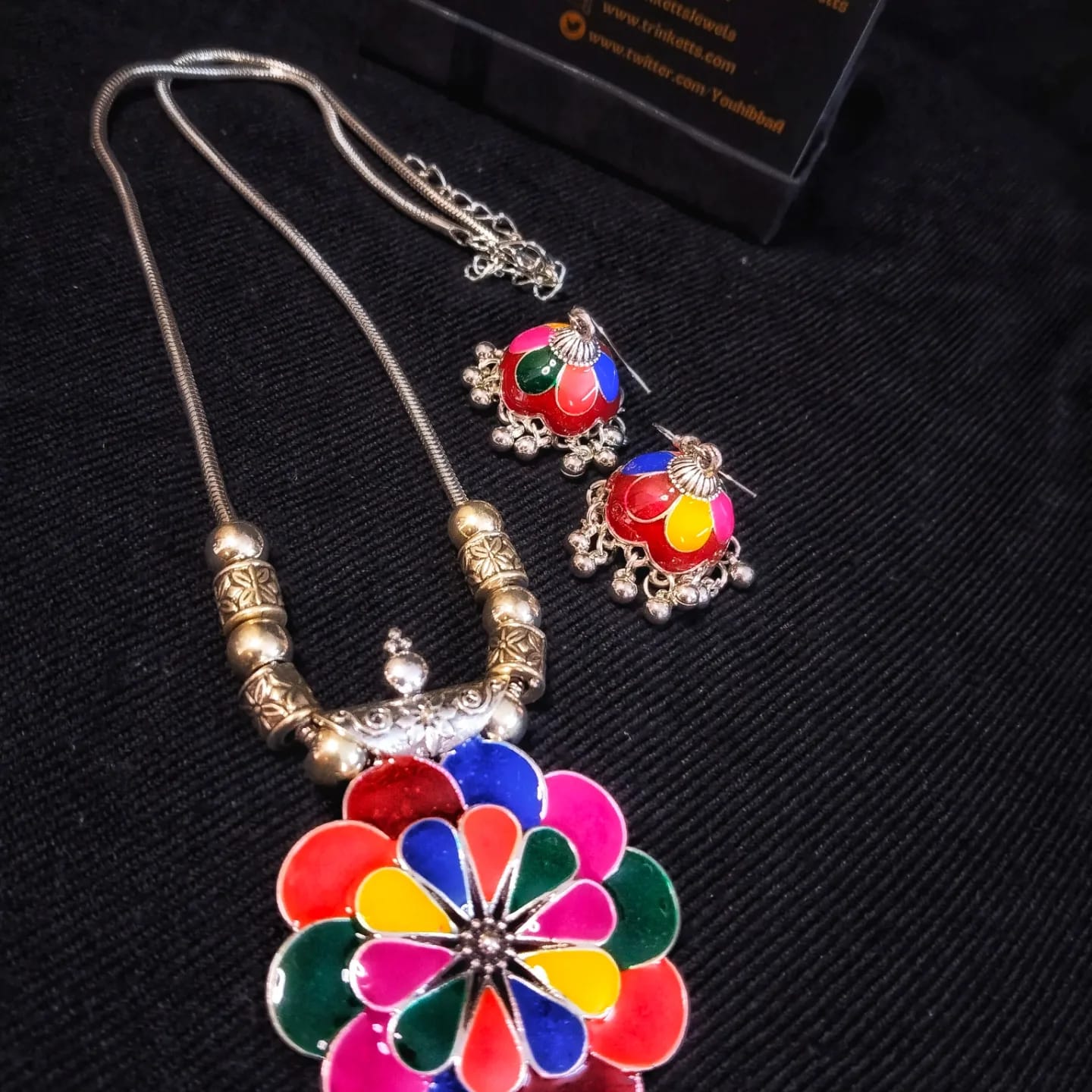 A floral-shaped pendant in multi colours Meena Kaari design, accompanied by matching Meena Karai jhumkis (dangling earrings) in multi colours, featuring attached ghungroos (small bells). The pendant hangs on an oxidized silver chain with an S hook closure.