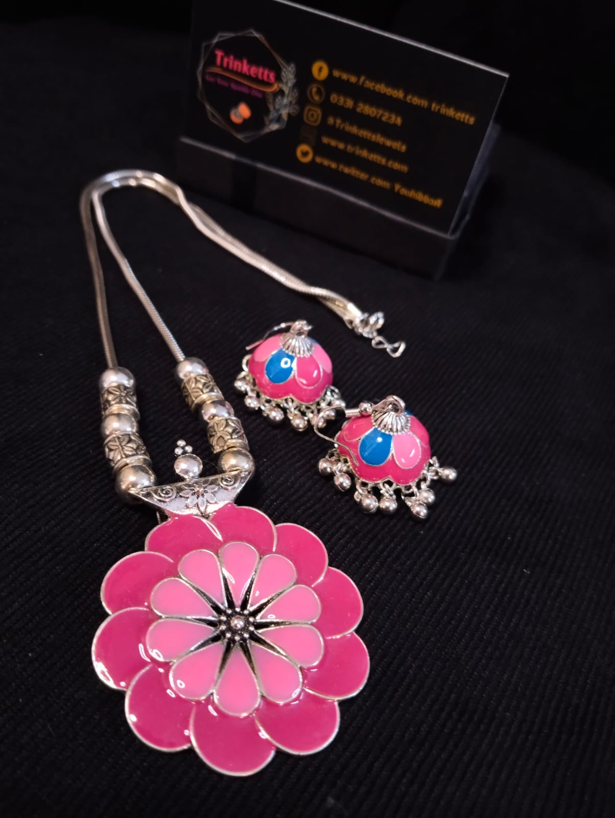 A floral-shaped pendant in pink and light pink Meena Kaari design, accompanied by matching Meena Karai jhumkis (dangling earrings) in in pink,light pink and blue, featuring attached ghungroos (small bells). The pendant hangs on an oxidized silver chain with an S hook closure.