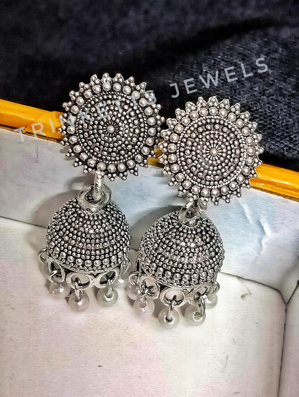 Oxidized silver metal Jhumki earrings, approximately 1.5 inches in size. The design features an intricate circular pattern with a hanging Jhumki plate adorned with white beads. Elegant and timeless, these Jhumkis add a touch of Indian charm to your style